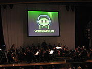 A photograph of an orchestra on a dimly lit stage. Above the group is a projection screen with a black, white, and green image of pixel art. The pixel art is of an oval object wearing headphones with eyes and four tentacles. Below the pixel art is the phrase