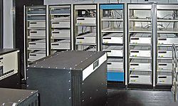 A room with a black box in the foreground and six control cabinets with space for five to six racks each. Most, but not all, of the cabinets are filled with white boxes.