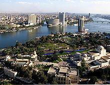 A sharp blue river divides the cityscape, which is primarily greenery and sparse low-rise buildings in the foreground, and dense with several modern high-rises in the background