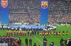 Two teams of sportsmen, one in yellow shirts and one in red and blue shirts, stand in a line. In front of the line is a line of children in equivalent shirts, but each in front of the opposite team. A number of other people stand in the foreground looking on. Behind the teams, a number of people hold a large circular piece of fabric with logos printed on, and two large banners hang down vertically from above the whole scene.